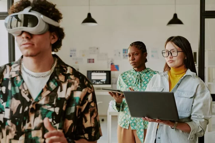 Two female developers observing a man using VR goggles in an office.